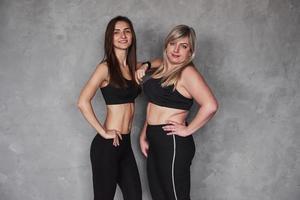 jung und erwachsen. Zwei Freundinnen mit unterschiedlichen Körpertypen und in Sportbekleidung stehen im Studio mit grauem Hintergrund foto