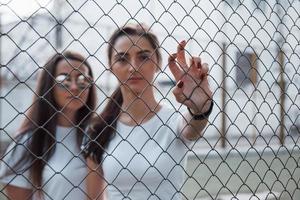 moderne Jugend. Zwei Freundinnen stehen tagsüber im Freien hinter dem Zaun foto