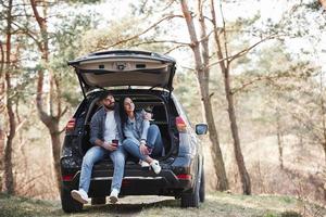 ländliche Szene. Sitzen auf dem hinteren Teil des Autos. die Natur genießen. Paar ist mit ihrem brandneuen schwarzen Auto im Wald angekommen foto