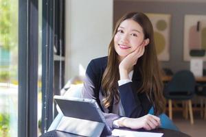 asiatische berufstätige frau in einem schwarzen anzug arbeitet an einem tablet auf dem tisch, lächelt glücklich im büro und arbeitet zu hause.