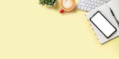 Büroschreibtisch mit Tastaturcomputer, Smartphone mit leerem Bildschirm, Tasse Kaffee, Stift, Notizbuch auf gelbem Hintergrund, Draufsicht mit Kopierraum, Attrappe. foto