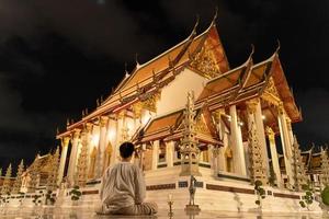 asiatischer buddhistischer mann in traditioneller thailändischer kleidung, der zur meditation sitzt, um der buddha-statue im wat suthat thepwararam am buddhistischen heiligen tag, dem buddhistischen sabbattag, achtung zu zollen, foto