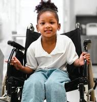 porträt eines süßen afroamerikanischen kindes, das lächelnd im rollstuhl im krankenzimmer sitzt. Kleines Mädchen kommt zur professionellen medizinischen Behandlung in die Klinik. Rehabilitationskonzept foto