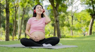 Schwangere Frau, die draußen im Park sitzt und ihren dicken Bauch mit roten Kopfhörern berührt, lässt ihr Baby beruhigende Harmoniemusik hören. schwangerschaftsreise, mutterschafts- und fötusentwicklungskonzept. foto