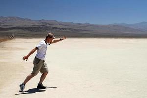 junger Mann mit Sonnenbrille und Shorts winkt mit den Armen, als stünde er mitten im Death Valley auf einem Surfbrett foto