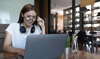 lächelnde asiatische Studentin im Grundstudium in der Bibliothek mit Laptop-Büchern, die Online-Recherchen für Kursarbeiten durchführen, Notizen für Aufsatz-Hausaufgaben machen, Online-Bildungs-E-Learning-Konzept foto