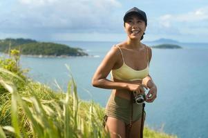 schöne asiatische frau in sportbekleidung, die nach dem trekking-, reise- und ökotourismuskonzept auf dem berggipfel am meer fotografiert. foto