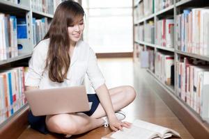 asiatische schöne Studentin, die in Bibliothek mit Laptop studiert foto
