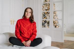 Foto einer angenehm aussehenden jungen Frau in rotem Pullover, Hose, sitzt mit gekreuzten Beinen auf einer bequemen Couch, genießt häusliche Atmosphäre, geschmückter Neujahrsbaum im Hintergrund. Home-Konzept