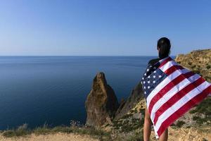 Rückansicht einer jungen Frau, die eine amerikanische Flagge hält, die an der Küste gegen das sonnige, helle Meer weht. foto