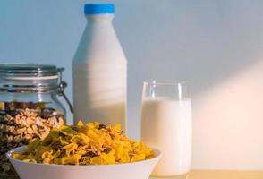 ein glas milch und milchflasche mit leerem etikett auf holztisch in der nähe einer schüssel mit müsli mit löffel. Kalziumnahrungsfrühstück für Kinder, bevor sie morgens zur Schule gehen. Cornflakes und Milchkonzept foto