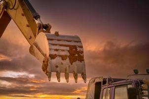 Schmutzmetalleimer des Baggers nach dem Graben des Bodens. Bagger geparkt in der Nähe von LKW mit goldenem Sonnenuntergang Himmelshintergrund. Raupenbagger. Erdbewegungsmaschine auf der Baustelle in der Abenddämmerung. Aushubfahrzeug. foto