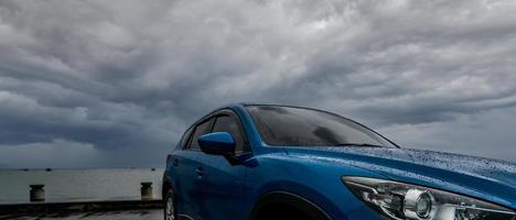 Blaues SUV-Auto mit Wassertropfen, das auf dem Parkplatz in der Nähe des Meeresstrandes gegen stürmischen und bewölkten Himmel geparkt ist. Vorderansicht des neuen Luxus-SUV-Autos mit Sportdesign. Mietwagen für Roadtrip. Autofahren an regnerischen Tagen. foto