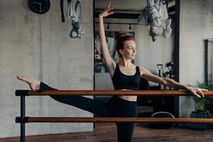 junge athletische rothaarige ballerina, die sich im fitnessstudio das bein auf der ballettstange dehnt foto