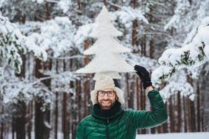 lustiger bärtiger mann mit bart, in warme winterkleidung gekleidet, hält künstlichen tannenbaum auf dem kopf, posiert gegen schneebedeckte bäume und ist gut gelaunt. Optimistischer Mann verbringt Zeit im Freien foto