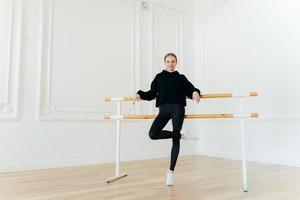 positive schlanke frau hat tanzunterricht, übt im studio in der nähe der ballettstange, steht auf einem bein, trägt schwarzes outfit und weiße sportschuhe, hat starke muskeln, schöne körperform. gymnastisches Konzept foto