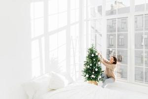 winterferien, neujahrskonzept. zufriedene junge brünette frau hat festliche stimmung, schmückt den weihnachtsbaum zu hause, bereitet sich auf die feier vor, posiert in einem gemütlichen schlafzimmer mit weißen wänden auf der fensterbank. foto