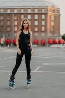 vertikale aufnahme einer gesunden schlanken frau, die in aktiver kleidung gekleidet rollschuh fährt, verbringt ihre freizeit im freien, lächelt glücklich und hat sportliche aktivitäten, die einen sportlichen lebensstil führen. Inline-Skating-Konzept foto