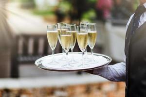 nicht erkennbarer kellner in formeller kleidung hält tablett mit champagnergläsern, trägt getränke auf festlichem tisch zu gästen, bietet guten service für besucher. Hospitality-, Service- und Trinkkonzept foto