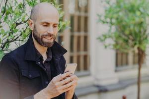 Ehrliche Aufnahme eines kahlen bärtigen Mannes mittleren Alters hat ein positives Lächeln, schaut aufmerksam auf das Smartphone, liest die notwendigen Informationen im Internet, posiert vor verschwommenem Hintergrund mit grünen Bäumen. foto