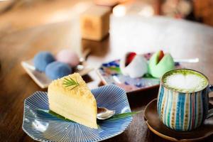 Nahaufnahme Frischkäse-Chiffon-Kuchen in einem gekachelten Teller, dekoriert mit Rosmarinblättern und Matcha-Grüntee auf der Oberseite dekorierte Latte Art mit herzförmigem Milchschaum in einer bunten Tasse, Kuchen im japanischen Stil foto