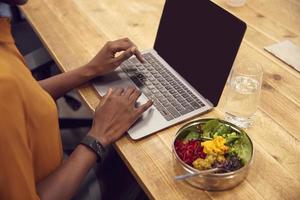 Geschäftsfrau, die am Laptop arbeitet, während sie veganes Mittagessen am Holztisch isst
