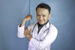 Ein Porträt eines jungen asiatischen männlichen Arztes, der seinen Bizeps isoliert durch einen blauen Hintergrund zeigt foto