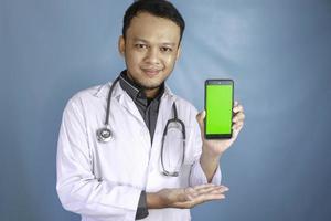 Der junge asiatische Arzt zeigt einen grünen Bildschirm oder Kopierbereich auf seinem Smartphone foto