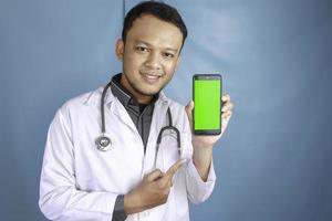 Der junge asiatische Arzt zeigt einen grünen Bildschirm oder Kopierbereich auf seinem Smartphone foto