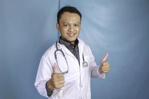 Porträt eines jungen asiatischen Arztes, ein Mediziner lächelt und zeigt Daumen hoch oder OK-Zeichen isoliert auf blauem Hintergrund foto