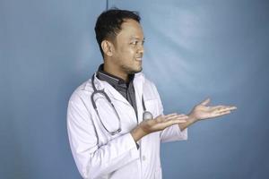 glücklicher junger asiatischer arzt, mediziner lächelt und zeigt auf einen kopierraum, der über blauem hintergrund isoliert ist foto