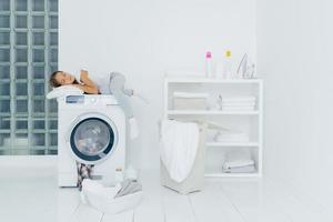 Vorschulkind schläft auf Waschmaschine, müde vom Waschen, posiert in weißer großer Waschküche mit Korb und Becken voller schmutziger Kleiderflaschen mit flüssigem Pulver. Kindheit, Hausarbeit