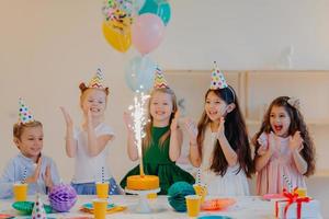 überglückliche kinderfreunde klatschen in die hände und schauen auf den kuchen, stehen in der nähe eines festlichen tisches mit geschenken, tassen und feiertagsattributen, haben gemeinsam spaß, feiern geburtstag foto