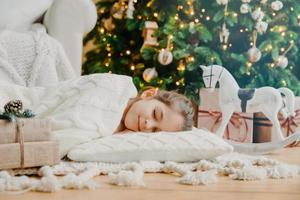 charmantes mädchen schläft auf weichem weißen kissen auf dem boden vor geschmücktem neujahrsbaum, hat angenehme träume, umgeben von spielzeugpferd und geschenkboxen. kinder-, rest- und winterferienkonzept. foto