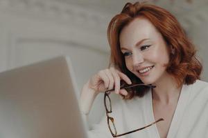 professionelle lächelnde rothaarige webdesignerin verwendet anwendung auf laptop-computer zum erstellen von projektarbeiten, recherchiert online und surft im internet, hält brillen in der hand, hat einen glücklichen ausdruck foto