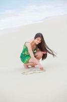 schönes jugendlich Mädchen, das Herz im Sand am tropischen Strand zeichnet foto