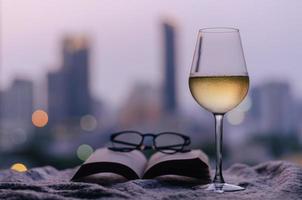 ein glas weißwein mit buch und brille auf dem bett mit buntem stadt-bokeh-lichthintergrund. konzept zu hause bleiben. foto