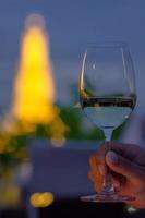 Hand, die nachts ein Glas Weißwein mit verschwommenem buntem Hintergrund des Tempels röstet. foto