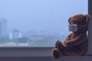 Teddybär mit Maske sitzt am Fenster, während es in der Monsunzeit regnet. foto