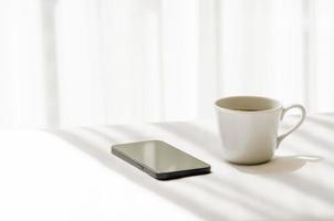 selektiver fokus einer tasse schwarzen kaffees und eines smartphones, die morgens auf ein weißes bettlaken mit schatten des vorhangs gelegt werden. entspannen zu hause konzept. foto