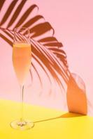 Ein Glas kalter Champagner mit Dampf, der Kokosblattschatten aus Sonnenlicht auf rosa und gelbem Hintergrund hat. Sommerkonzept. foto