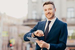 aufnahme eines gutaussehenden männlichen unternehmers trifft sich mit geschäftsleuten, schaut auf armbanduhr, verwendet touchpad für die suche nach notwendigen informationen im internet, trägt eleganten formellen anzug und krawatte foto