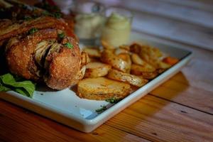 deutsche schweinshaxe wird mit gebackenen kartoffeln und sauce in einem weißen keramikteller serviert, der auf dem esszimmertisch platziert wird, um die schweinshaxe bereit für ein festliches abendessen zu machen. foto