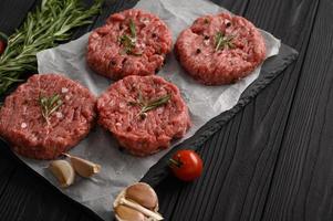 Raw Beef Burger Patties auf Metzgerholzbrett, Rosmarin und Pfeffer. schwarzer Hintergrund. Sicht von oben. foto