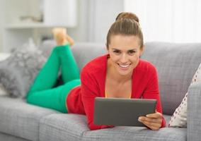 lächelnde junge Frau, die auf Couch mit Tablette PC liegt foto