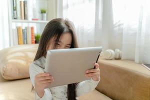 süßes Kind, das ein Tablet benutzt und lächelt, während es zu Hause auf dem Sofa sitzt foto