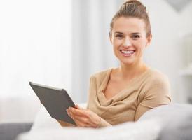 lächelnde junge Frau, die auf Sofa sitzt und Tablet-PC verwendet foto