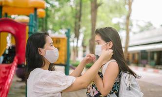 mutter legt eine schutzmaske auf das gesicht der tochter zum schutz von covid-19 oder coronavirus-ausbruch im dorfpark, um sich auf den schulbesuch vorzubereiten. zurück zum schulkonzept. medizinische maske zur vorbeugung von coronavirus. foto