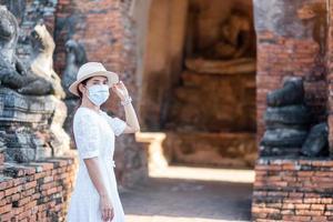 touristische frau in weißem kleid mit chirurgischer gesichtsmaske, schutz covid-19 pandemie während des besuchs im wat chaiwatthanaram tempel in ayutthaya. neues normal-, sicherheitsreise- und thailand-reisekonzept foto