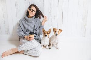 glückliche brünette frau in lockerer hauskleidung gekleidet, froh, sitzt auf dem boden, entspannt sich mit heißem cappuccino in pappbecher, verbringt zeit mit hunden, isoliert über weißem zaunhintergrund foto